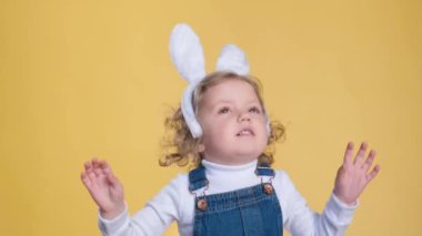 Tavşan kulağı ve tulum giyen mutlu bir çocuk sarı bir arka planda duruyor, baş parmağıyla gülümsüyor ve el sallıyor, neşe ve neşeyi neşeli bir parmakla paylaşıyor..