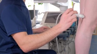 Doktor hastaların bacağını muayene etmek için ultrason makinesi kullanıyor. El ve bilekleriyle jestler yapıyor..