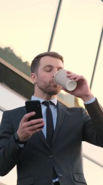 Sakallı ve takım elbiseli bir adam, kahvesini yudumluyor, telefonuna bakıyor, parmağını oynatıyor, başparmağını yakasına yaklaştırıyor..