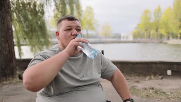 疲れた若い太った男は 屋外に座っている間 ボトルから水を飲んで灰色のTシャツを着た トレーニング中に休憩する 壊れてる 一時停止する — ストック動画