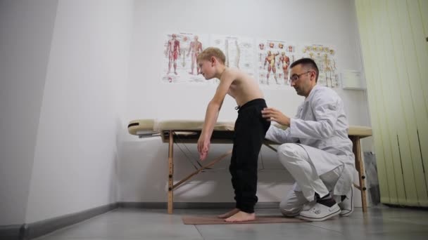 病院では 医者が木製の椅子に座っている間 若い男の子の足を評価しています — ストック動画
