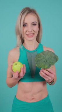 Kadın mutlu bir şekilde elinde bir elma ve yüzünde bir gülümsemeyle brokoli tutuyor..