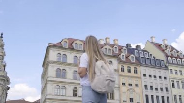 Sırt çantalı bir kadın bir binanın önünde duruyor, etkileyici dış görünüşüne bakıyor, canlı şehir manzarasıyla çevrili..