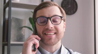 Gözlüklü ve gülümseyen bir doktor cep telefonuyla konuşuyor, kendinden emin bir jest yapıyor..