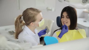 Gözlük takan bir kadın dişçi dişlerini incelerken bir diş sandalyesinde oturur. Boş zamanlarında gülümser ve mutlu bir şekilde el kol hareketi yapar..