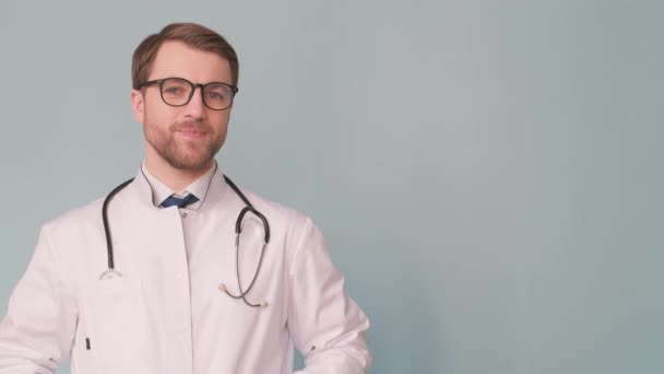 一位戴眼镜 脖子上戴听诊器的医生站在一面蓝色的墙上 脸上挂着笑容 — 图库视频影像