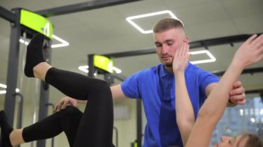 Bir erkek spor salonunda bir kadına egzersizlerde yardım ediyor, jestlerle eklem ve kas hareketlerine odaklanıyor ve spor ekipmanları ve spor kıyafetleri kullanıyor..