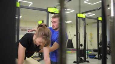 Bir erkek spor salonunda egzersiz yapan bir kadına yardım ediyor. Eşyalar ve tişörtlerle dolu bir odada beden eğitimi tekniklerini paylaşıyor..