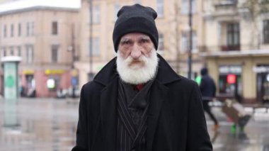 Şehrin sokaklarında sakallı yaşlı bir adamın yakın plan portresi, buruşuk yüzü. Düşünceli, üzgün yaşlı adam kameraya bakıyor..
