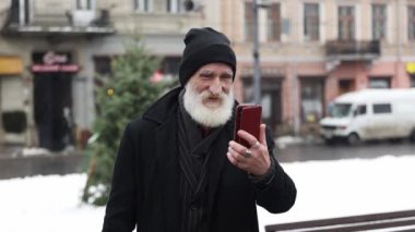 Siyah kıyafetli yaşlı, kır saçlı bir büyükbaba kış sokağının ortasında dikiliyor ve bir video görüşmesinde konuşuyor. Mutlu büyükbaba telefonda konuşuyor.