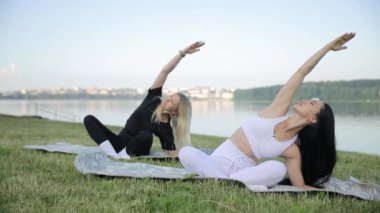 İki genç kadın göl kıyısında sabahları yoga yapıyor, gövdelerini büküyorlar. Sağlık konsepti. Duygusal ve fiziksel sağlığa dikkat edin.