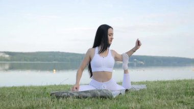 Güzel, uzun saçlı bir esmer göl kıyısında yoga yapıyor. Duygusal ve fiziksel sağlığa dikkat edin.