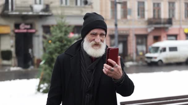 一位穿着黑色外套的白发苍苍的老爷爷站在一条过冬的大街上 一边打电话一边说着话 爷爷打电话聊得很开心 — 图库视频影像