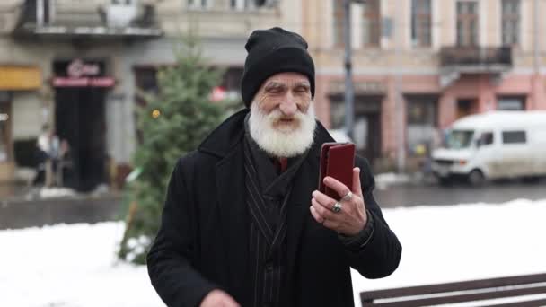 黒いアウターウェアのポジティブな祖父は 冬の外に立っている間にビデオ通話で話しています ハッピーおじいちゃんが電話で話して 誰かに手を振り 笑顔を浮かべました メリークリスマスオンライン — ストック動画