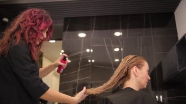 Bir kuaför, genç sarışın bir kadının saçını bir güzellik salonunda sandalyede otururken spreyler. Saç bakım konsepti. 