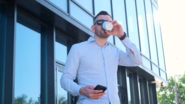 Resmi kıyafetli bir adam kahve içiyor ve cep telefonu kullanıyor. Sakalı ve güneş gözlüğü var. Yaz günü