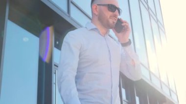 Siyah gözlüklü bir adam bir ofis binasının önünde cep telefonuyla konuşurken el kol hareketi yapıyor. Müdahaleciye karşı saldırganlık ve öfke gösteriyor gibi görünüyor.