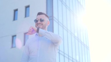 Sakallı ve güneş gözlüklü bir adam, geçmişinde mavi bir bina olan akıllı bir saat kullanarak iletişim kurar..