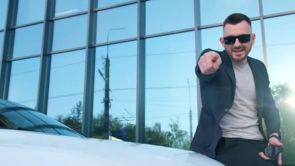 スーツとサングラスの男は 背景にオフィスビルを持つ車の隣に立っている間 カメラにジェスチャーしています — ストック動画