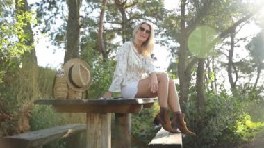 Ormandaki ağaçlarla çevrili ahşap bir masada oturan bir kadın, her yerde yemyeşil çimenler ve bitkiler var. Gözlük takarak manzaranın tadını çıkartıyor..