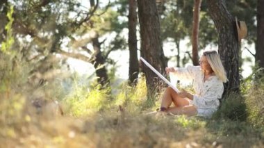 Bir kadın bir bitkinin altında oturmuş, doğal bir ormanlık alanda kitap okuyor. Ağaçlar ve dallar barışçıl bir atmosfer yaratırken çimenli orman zemini onu çevrelemektedir..