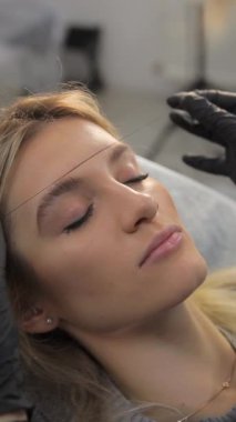 Bir kadın, kaşlarını güzellik salonunda yaptırırken kirpiklerini yaptırıyor ve muhtemelen burun, saç, yanak ve çene gibi diğer yüz hatlarını da yaptırıyor..