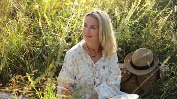 Kobieta Białej Sukience Kapeluszu Uśmiecha Się Siedząc Trawie Naturalnego Krajobrazu — Wideo stockowe