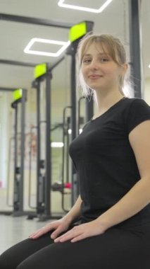 Siyah elbiseli, jimnastik minderinde oturan güzel bir genç kızın dikey videosu. Rehabilitasyon merkezinde bir kız. Fitness kızı.