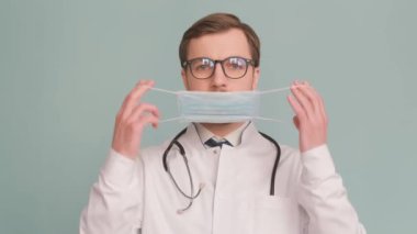 Gözlüklü ve steteskoplu bir doktor, yüzünde tıbbi bir maske tutuyor, gülümseme ve endişenin jestleri sırasında görme bakımının önemini gösteriyor..