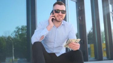 Sakallı bir adam bir binanın merdivenlerinde oturuyor güneş gözlüğü takıyor ve cep telefonuyla konuşurken para tutuyor. İş adamları