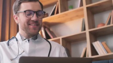 Yüzünde gülümseme olan bir doktor, gözlüklü ve boynunda steteskopla tablet bilgisayar kullanıyor..