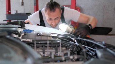Bir otomotiv hizmet binasında el feneri kullanarak bir araç motoru üzerinde çalışan bir tamirci, motorlu araçların makine bileşenlerine odaklanıyor.