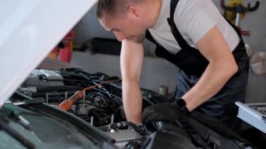 Şapka takan bir adam garajda bir aracın kaputunun altında çalışıyor. Otomotiv lastiğini tamir ediyor. Silecekleri ve ön camı kontrol ediyor..