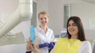 Bir kadın dişçinin yanındaki dişçi koltuğunda oturuyor, saçları düzgünce düzenlenmiş ve yüzünde hoş bir gülümseme var..