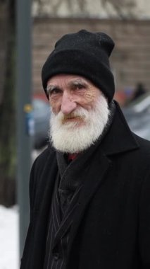 Kameraya bakan kalın gri sakallı yaşlı bir adamın yakın plan portresi. Şehirdeki ciddi, dalgın, kırışık büyükbaba.