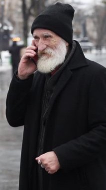 Yaşlı bir adamın kışın dışarıda dikilirken telefonla konuşmasının dikey video portresi. Siyahlı yalnız bir emekli telefonda biriyle konuşuyor.
