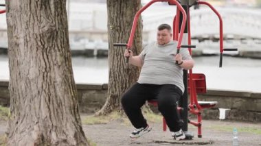 Gri tişörtlü genç şişman bir adam güpegündüz yeşil bir alanda egzersiz makineleriyle çalışıyor. Adam kilo veriyor. Fazla kiloyla mücadele et.