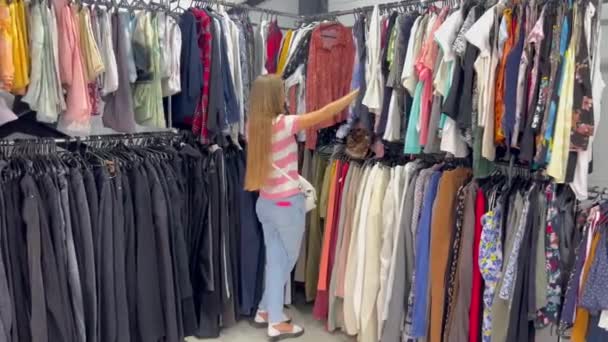 女性はショートパンツ Tシャツ スポーツウェアを含む服の小売店を閲覧し さまざまな色とデザインの服のハンガーのアイテムを調べています — ストック動画