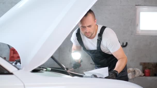 男はガレージで車を操作し フードを開けた自動車の外装に取り組んでいる — ストック動画