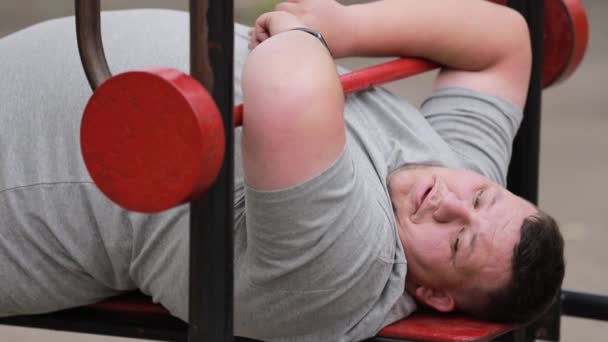 エクササイズマシンで休んでいる太りすぎの男性のクローズアップ 屋外の運動機械で重量を持ち上げる若い脂肪の男 — ストック動画
