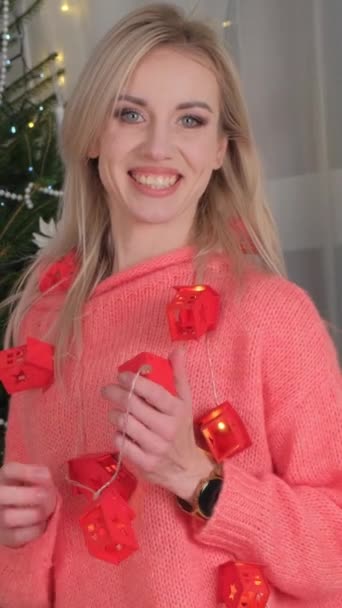 彼女の顔に笑顔でピンクのセーターで幸せなブロンドの女性は クリスマスツリーの前にワインのグラスを保持し お祝いのイベントに楽しいタッチを追加 — ストック動画