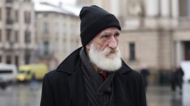 Şehrin sokaklarında sakallı yaşlı bir adamın yakın plan portresi, buruşuk yüzü. Düşünceli, üzgün yaşlı adam kameraya bakıyor..