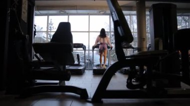 Bir kadın, cam pencereli ve ahşap döşemeli bir binada spor salonunda koşu bandında egzersiz yapıyor..