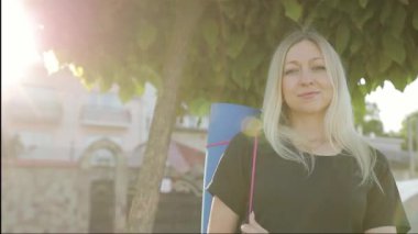 Güzel orta yaşlı bir kadın sırtına jimnastik minderi tutarken kameraya bakıyor. Eğitimden önce. Güneş ışığının arka planında ağacın altında duran fitness kadını.