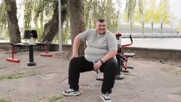 満足している太りすぎの若者は公園のエクササイズマシンに乗っている 野外運動機の近くの緑地に灰色のTシャツを着た太った男 トレーニング後の疲労 — ストック動画