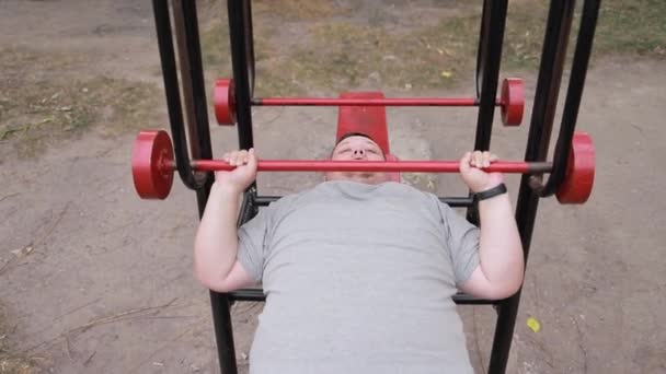 永続的な脂肪の男は屋外の運動機械で横たわっているウェイトを持ち上げます 過剰な体重で戦っている アスファルトの背景のトレーニングマン — ストック動画