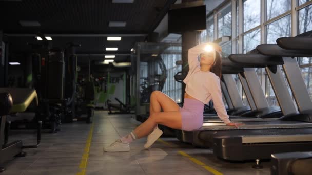 在体育馆里 一个穿着运动服的女人坐在跑步机上 在硬木地板上锻炼她的大腿和膝盖 — 图库视频影像