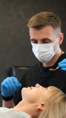 Bir dişçi, maske takıyor, ağzında mavi eldivenlerle bir kadının dişlerini inceliyor..