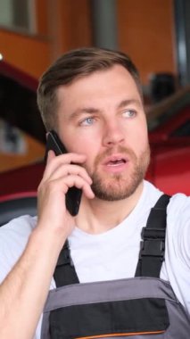Sakallı ve sakallı bir adam kırmızı bir arabanın önünde başparmağıyla el kol hareketi yaparken cep telefonuyla konuşuyor..