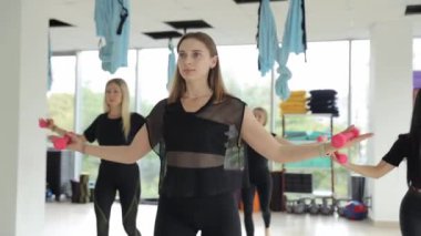 Spor salonundaki bir grup kadın dambıllarla egzersiz yapıyor, eklem, omuz ve bacak hareketlerine odaklanıyor. Yüzlerindeki gülümseme olaya duydukları heyecanı yansıtıyor..